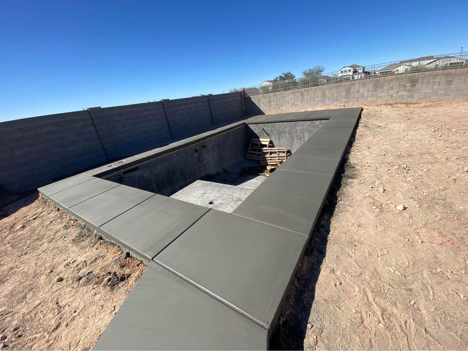 A rectangular swimming pool is under construction, showcasing a specialized installation with freshly laid concrete edges. Wooden pallets rest inside the empty pool against a backdrop of clear blue sky. A tall stone fence borders the area, highlighting concrete expertise in every detail.