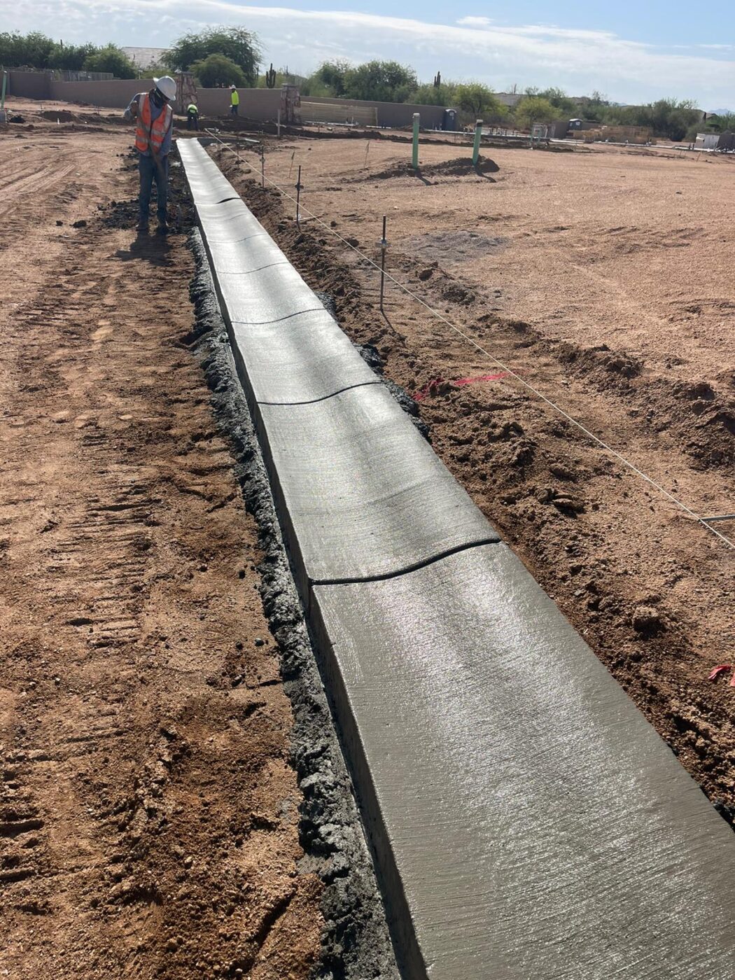 A licensed concrete contractor stands next to a freshly poured concrete curb under a clear sky. The long, smooth curb stretches through the sandy construction site. Equipment and other workers are visible in the background, showcasing professional concrete services in action.
